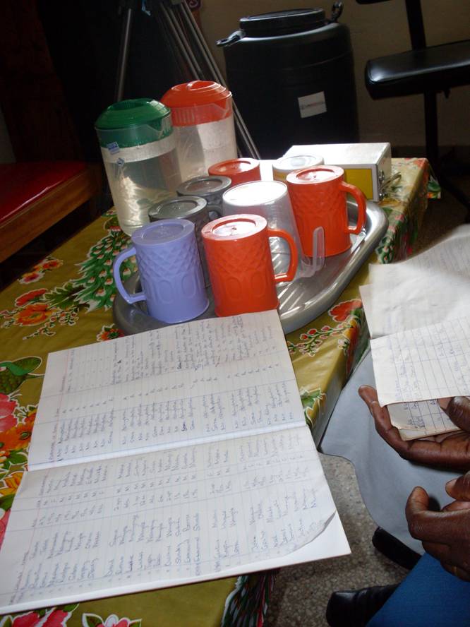 A table with a colorful tablecloth holds a tray with several cups and containers. In the forefront, open notebooks with handwritten notes are visible, and a person's hand is holding a piece of paper.