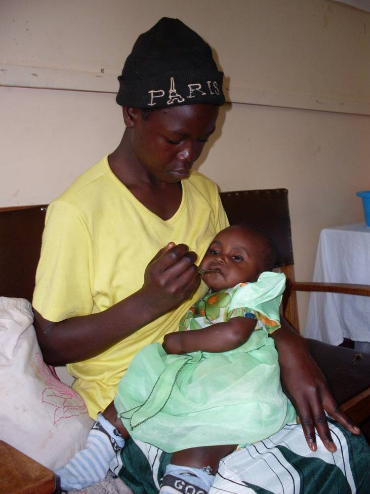 A person wearing a yellow shirt and a black beanie labeled "Paris" is sitting on a couch, feeding an infant dressed in a light green outfit. The setting appears to be indoors, with a pillow and a table in the background.