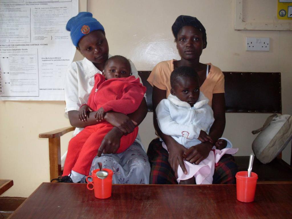 Two women sitting with two toddlers on their laps in a waiting room. The children are wearing red and white clothing. Two red cups are on the table in front of them. Medical posters are on the wall in the background.