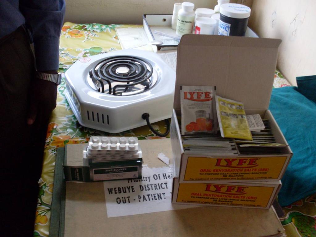 A table with various medical supplies, including a single burner electric stove, boxes of Lyfe oral rehydration salts (ORS), medication blister packs, and several bottles of medicine. A sign on a cardboard box reads "MINISTRY OF HEALTH WEBUYE DISTRICT OUT PATIENT.