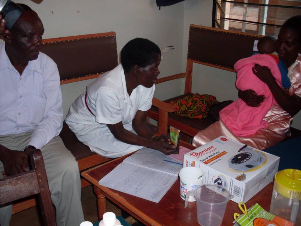 A healthcare worker in a white uniform sits with a man and a woman holding a baby. They are gathered in a clinic setting with medical supplies on the table, discussing or reviewing documents.