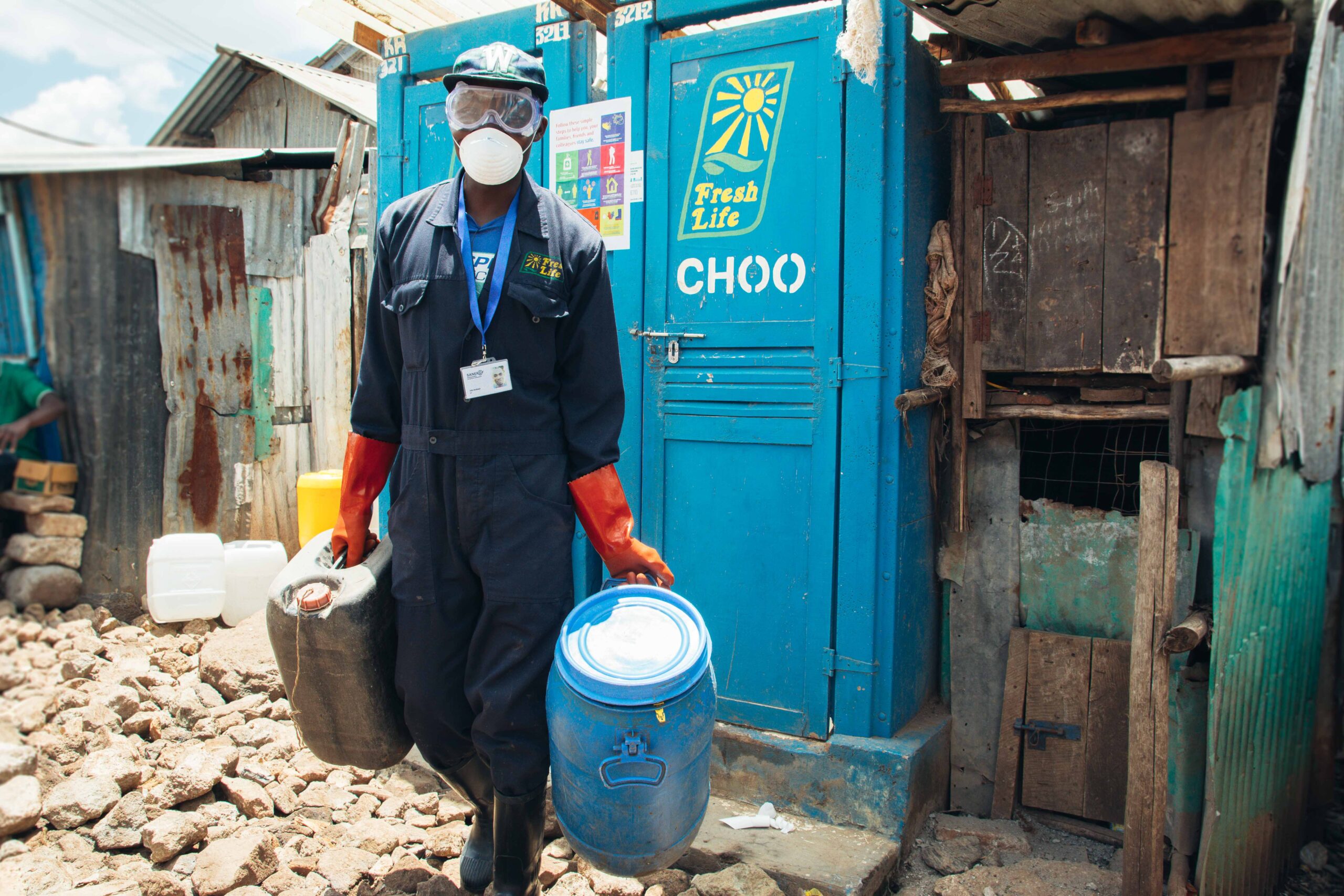 A person wearing a protective jumpsuit, mask, and gloves carries two containers, one blue and one black, outside a blue structure labeled "Fresh Life Choo" in an informal settlement area with rocky ground and makeshift buildings.