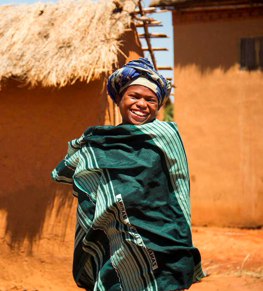 A person stands outside, smiling joyfully, wearing a headscarf and wrapped in a green and white striped fabric. The background includes earth-toned huts with thatched roofs, under a clear blue sky. The scene appears warm and sunny.