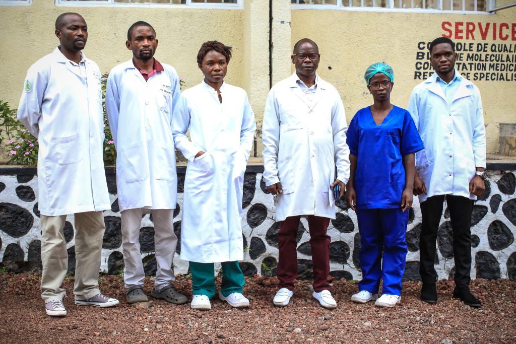 A group of six healthcare professionals, five wearing white lab coats and one wearing blue scrubs, stand in front of a beige building with a stone foundation. They face the camera directly, posing in a line. The wall behind them features written text.