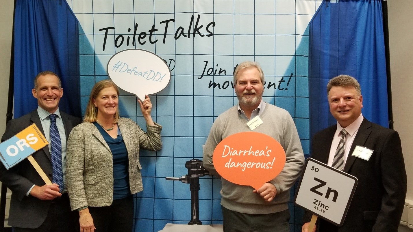 Four people are posing in front of a backdrop with the words "ToiletTalks" and "Join the movement!" (stylized as "#DefeatDD!"). They are holding speech bubbles and signs that read "Diarrhea's dangerous!," "ORS," "#DefeatDD!," and "Zn," suggesting a health-related event.