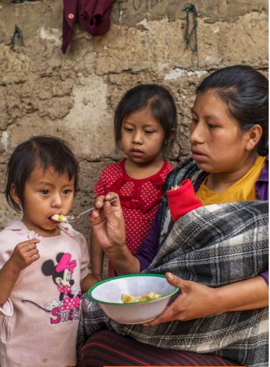 A woman holding a child in a blanket feeds a young girl with short hair a spoonful of food from a bowl. Another young girl stands nearby. They are indoors with a rustic wall in the background.