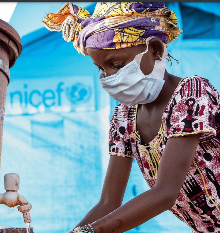 A young girl is washing her hands at a water tap. She is wearing a colorful headscarf and patterned dress, along with a face mask. The background is blue with partial text visible.