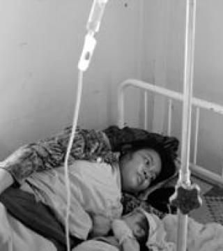 A person lies on a hospital bed with an IV drip attached, resting on their side and looking upwards. The room has minimal furnishings and a plain wall in the background.