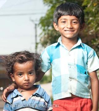 Two young boys are posing outdoors. The older boy, smiling and wearing a blue and white checkered shirt, has his arm around the younger boy, who has curly hair and a serious expression, wearing a blue striped shirt. They are standing on a sunny day. © PATH/Gabe Bienczycki.