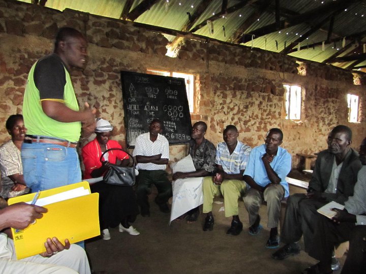 A group of people sit in a circle inside a stone building, engaged in discussion. One person stands, speaking. A chalkboard with writing is in the background. Some individuals hold papers, while others listen attentively. Natural light filters in through windows.