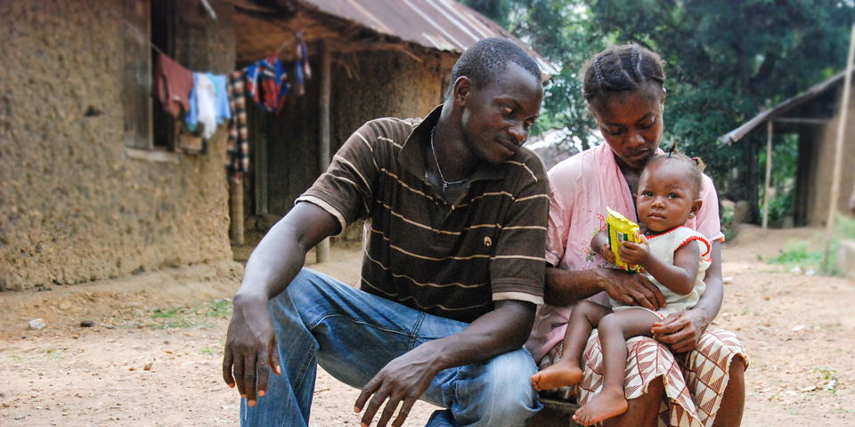 A man, woman, and baby sit outside a mud-brick house with a tin roof. The man, dressed in a striped polo and jeans, looks at the woman holding the baby. The woman, in a striped skirt and checked blouse, holds a packet while the baby sits on her lap, holding the packet.