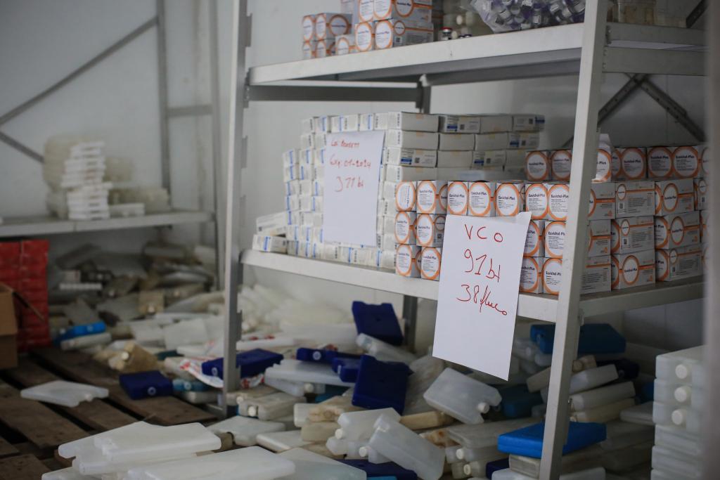 A storage room with metal shelves stocked with boxes of medical supplies. Two handwritten signs on the shelves indicate items labeled "VCO" and "Berber" with quantities and weights. The floor has various scattered items, including ice packs and foam padding.