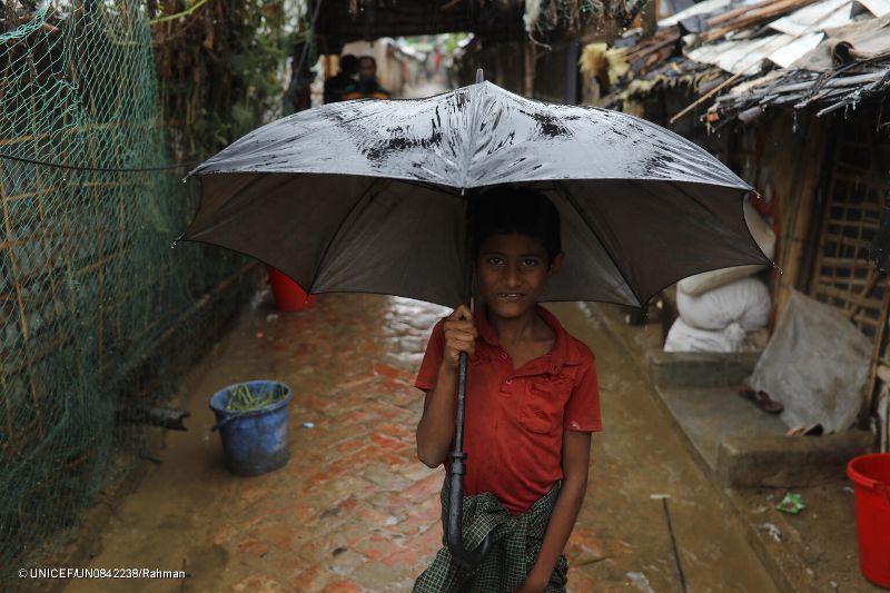 A young boy stands on a wet, narrow alleyway holding a large black umbrella. He wears a red shirt and a colorful cloth wrapped around his waist. The surroundings include makeshift shelters and a muddy ground, indicating a rainy day.