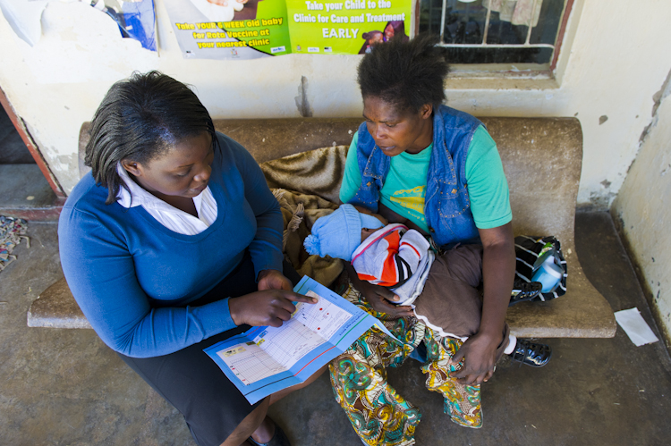 A health worker in a blue sweater sits on a bench explaining health information to a woman holding a baby wrapped in a blanket. They are in a clinic with informational posters on the wall behind them.