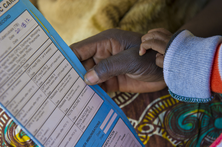 An adult hand holding a vaccination record booklet while a small child’s hand grasps the adult's thumb. The vaccination record is filled out with details. The background features a colorful, patterned fabric.