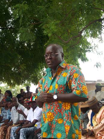 A man stands and speaks to a group of people seated outdoors under a tree. He wears a colorful shirt with a vibrant floral pattern and glasses. The audience listens attentively, some dressed in traditional attire.