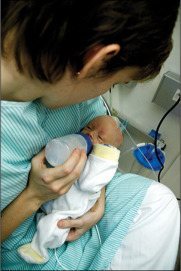 A person dressed in a hospital gown holds a newborn baby. The person is feeding the baby a bottle. The baby is wrapped in a white blanket, and medical equipment is visible in the background. The person leans in closely to the baby.