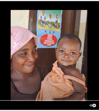 A woman in a pink headscarf smiles while holding a baby dressed in orange. Behind them, there is a poster on the wall showing several children and some text. The scene appears to be indoors with a wooden door in the background.