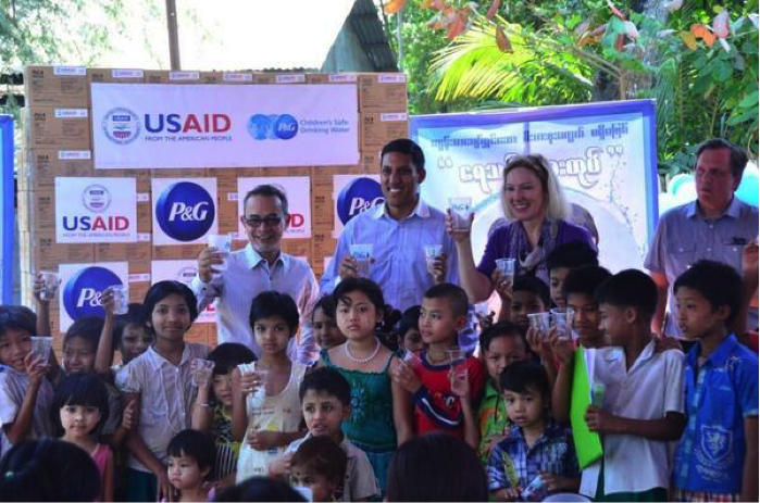 A group of adults and children gather outside in front of a display of USAID and P&G boxes. The adults hold up cups of water, smiling, while children stand and sit in front of them. Trees and a building are visible in the background.