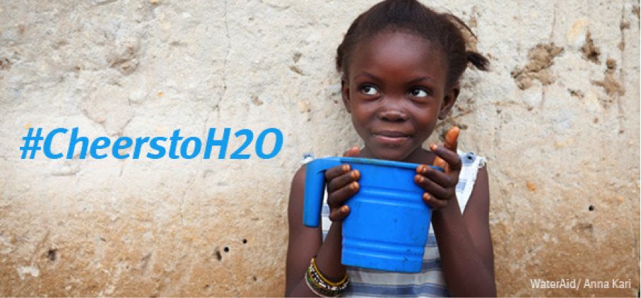 A young girl sits against a concrete wall, holding a blue container with both hands. She is smiling and looking to the side. The text "#CheerstoH2O" is on the left side of the image.