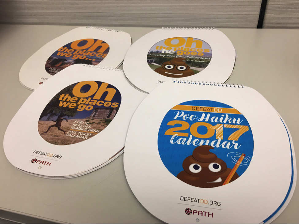 Spiral-bound calendars resembling toilet seats are arranged on a desk. The covers feature various titles, including "Oh the places we go," "Oh the places poo goes," and "Poo Haiku 2017 calendar." The calendars are associated with DEFEATDD.org and PATH.