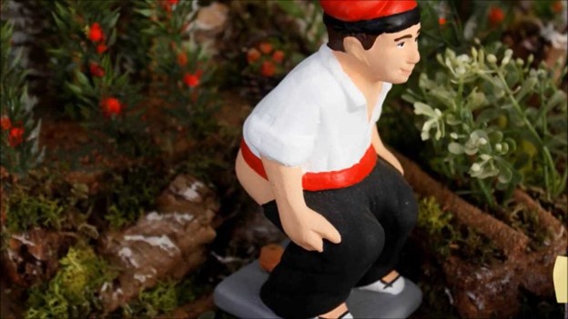 A figurine of a person in traditional attire with a red hat and sash, crouching on a flat surface. The background features greenery and small red berries.