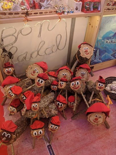 A display of traditional Catalan "Tió de Nadal" figures, crafted from logs with painted smiling faces and red hats, arranged on the floor. A sign above reads "Bon Nadal," meaning "Merry Christmas" in Catalan.