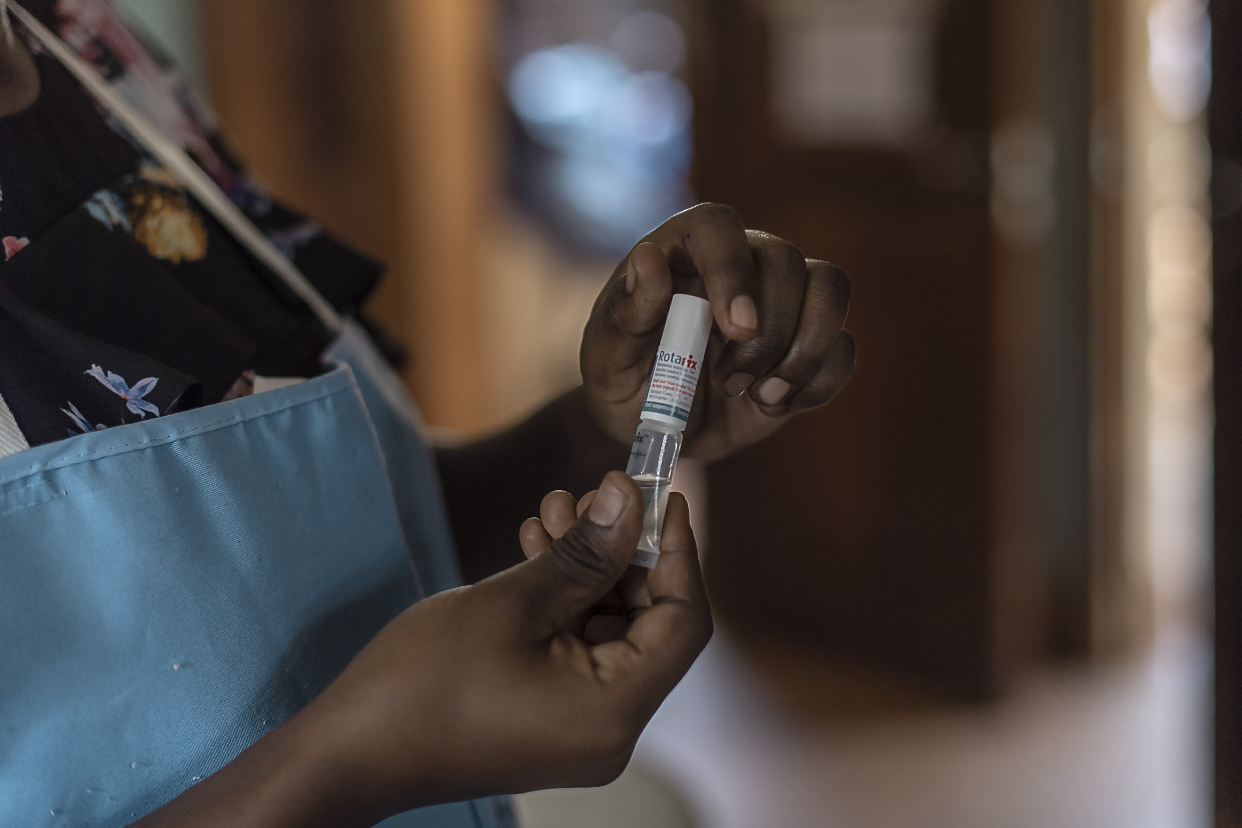 A person in a blue apron prepares a syringe, drawing liquid from a small vial indoors. The focus is on the hands and the vial. The background is softly blurred, with a door in the distance.