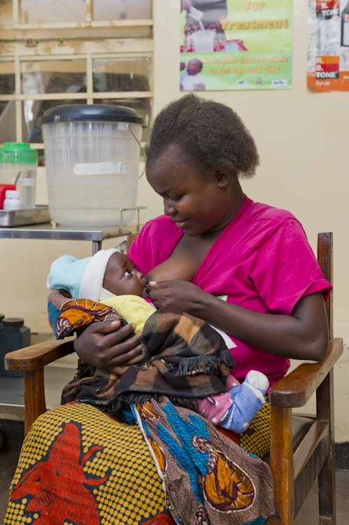 A woman in a pink shirt smiles while holding a baby wrapped in a blanket. They are sitting on a wooden chair inside a room with posters on the walls and medical equipment in the background.