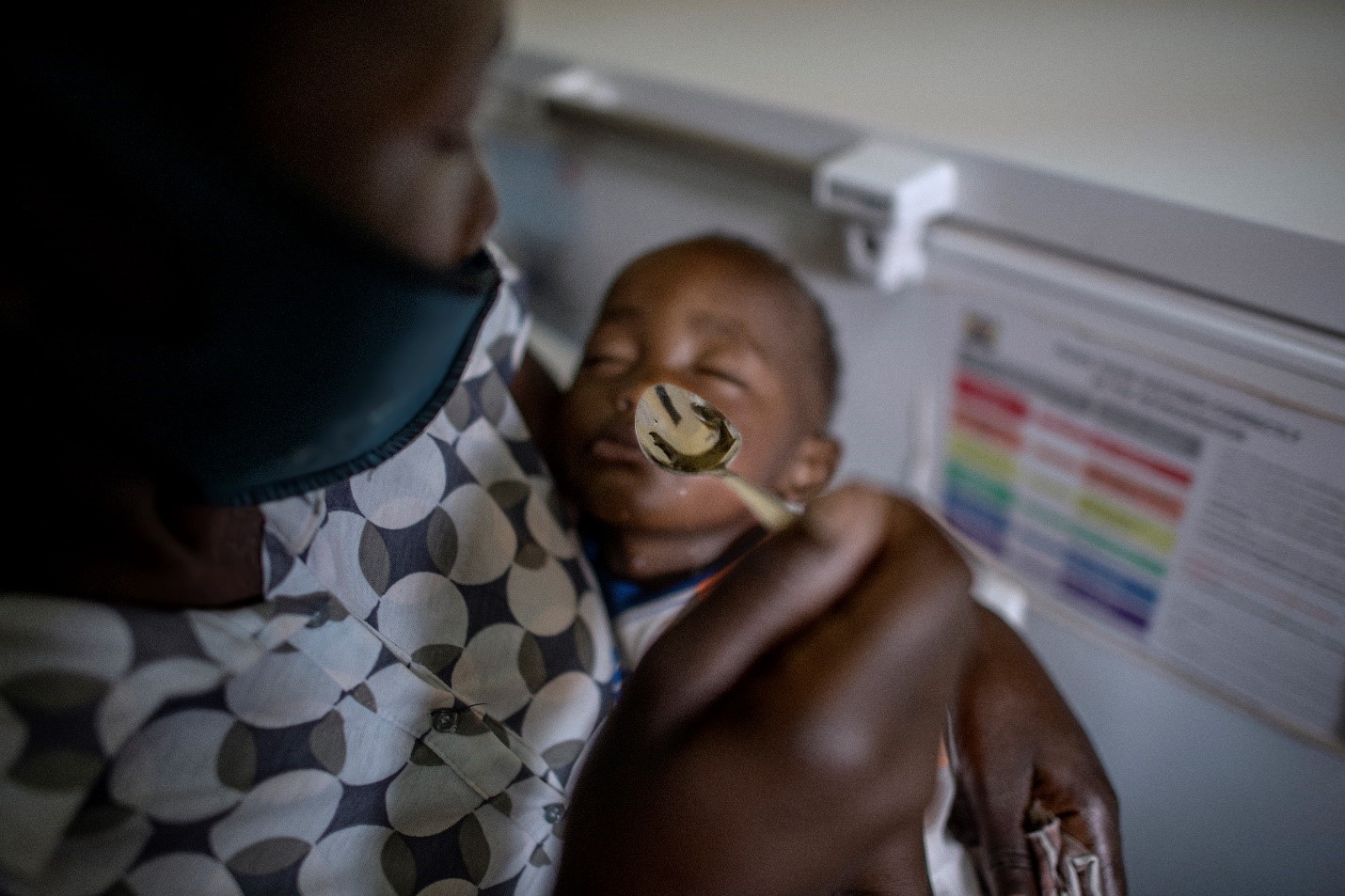 A caregiver spoons food into the mouth of a sleeping baby. The infant is cradled in the caregiver's arms, wearing a patterned shirt. The background is out of focus, featuring a chart with colorful sections.