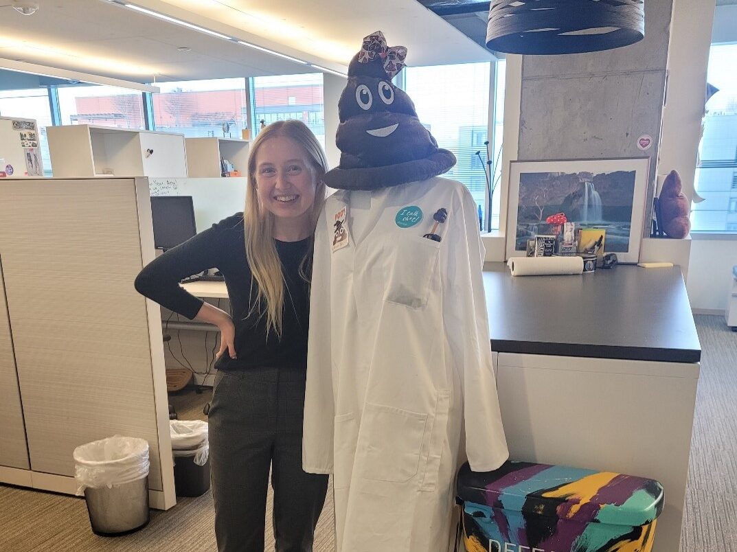 A woman with long blonde hair stands smiling next to a mannequin wearing a white lab coat and a poop emoji hat. The mannequin is positioned in an office space near colorful stools.