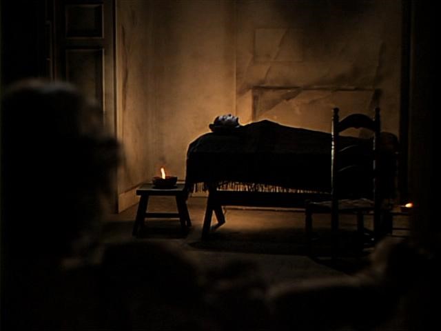 Dimly lit room with a bed covered by a dark cloth. A small table with a lit candle stands beside it. Shadows dominate the scene, creating a somber atmosphere.