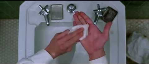 A person washing hands over a white sink with a bar of soap. The sink has two faucets and a small plug in the center. The background shows a tiled floor.