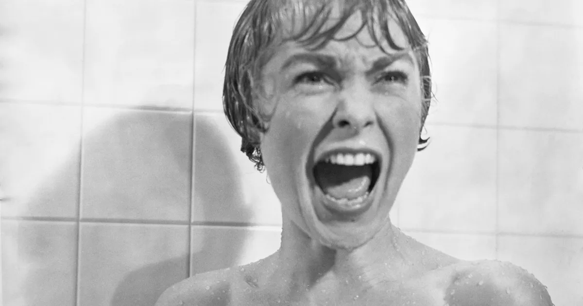 Black and white image of a woman with short, wet hair standing in a shower, screaming with a terrified expression. The background features tiled walls.