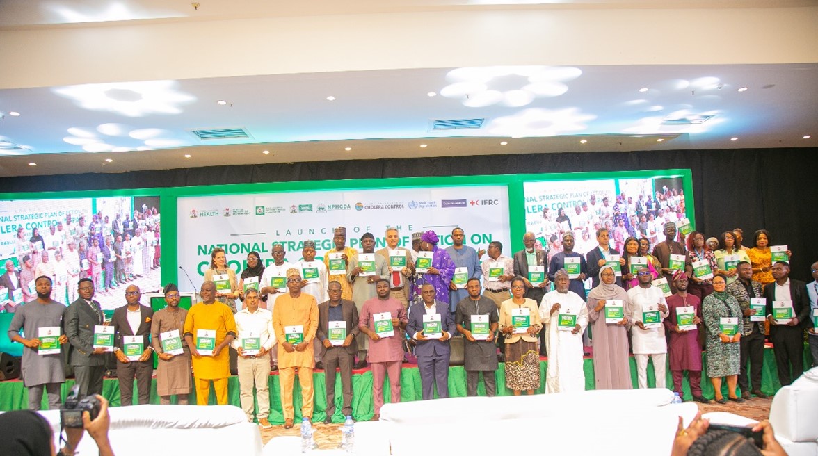 A group of individuals standing on a stage, each holding a booklet. They are posed in rows with a colorful backdrop displaying text related to Nigeria's National Strategic Plan of Action on Cholera Control. The scene is bright and well-lit.