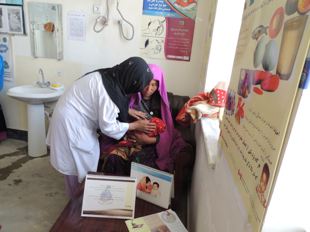 A healthcare worker in Afghanistan assists a woman in a purple headscarf who is breastfeeding her baby in a clinic room with health posters and educational materials on display.