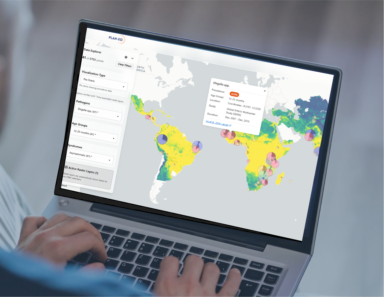 A person uses a laptop displaying a colorful world map with data overlays, pop-up information box, and filter options on the left side of the screen. The screen shows the Plan-EO dashboard, which uses big data to track diarrheal disease indicators.