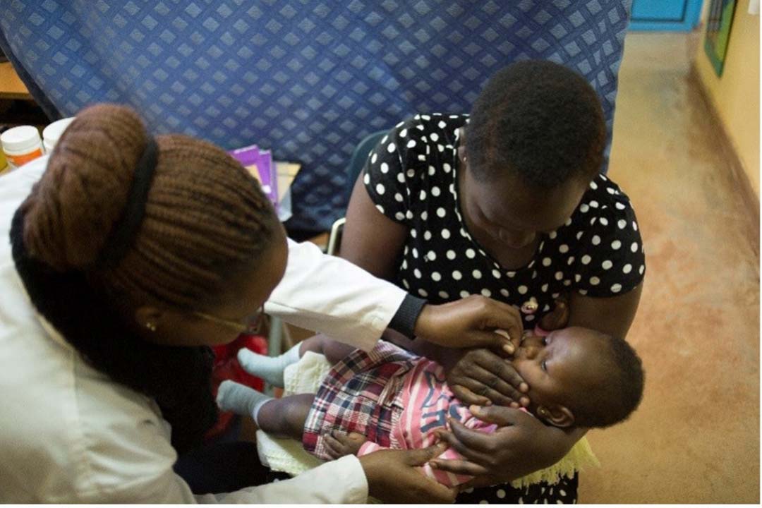 A healthcare worker in Kenya wearing a white coat gives an oral vaccine to a baby held by a woman in a black polka-dot dress, inside a clinic with a blue patterned curtain in the background.