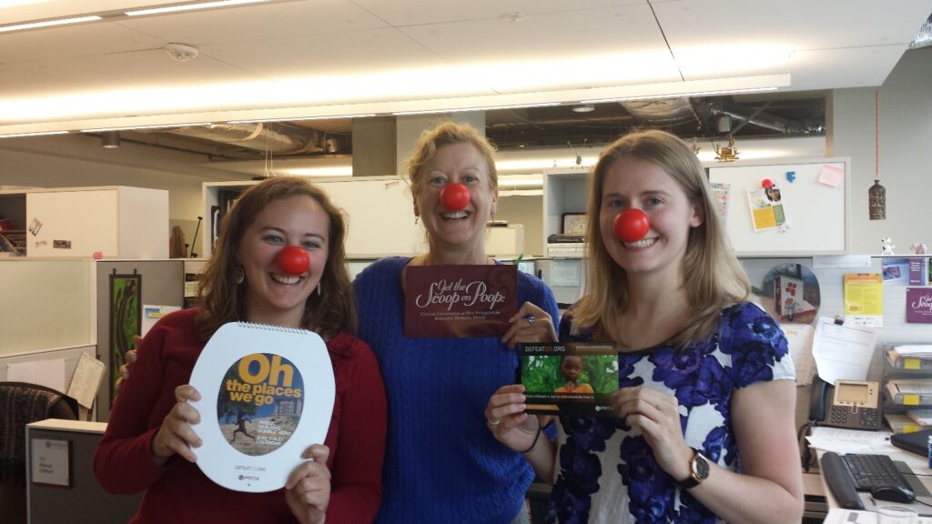 Three women wearing red clown noses stand in an office. One holds a sign that says “Oh the places we go,” another holds a card, and the third holds a postcard with a child’s photo. They are smiling and standing close together.