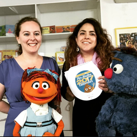Two women stand smiling with a girl Muppet puppet and a blue furry Muppet. One woman holds a calendar with a Muppet face on it. Shelves with books and Muppet merchandise are in the background.