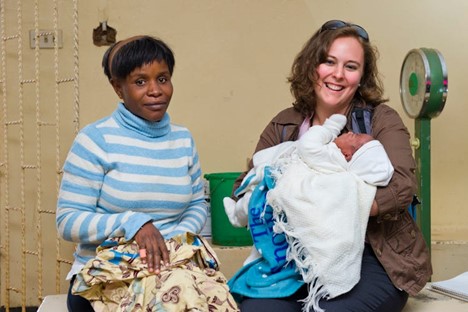 Two women sit side by side indoors; one holds a baby wrapped in white and blue blankets and smiles at the camera, while the other, wearing a striped sweater, looks toward the camera with a neutral expression. A scale is seen in the background.