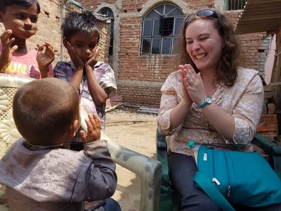 A woman sits outside smiling and clapping hands with a young boy, while two other children standing nearby also clap and smile. The background shows a brick building under construction.